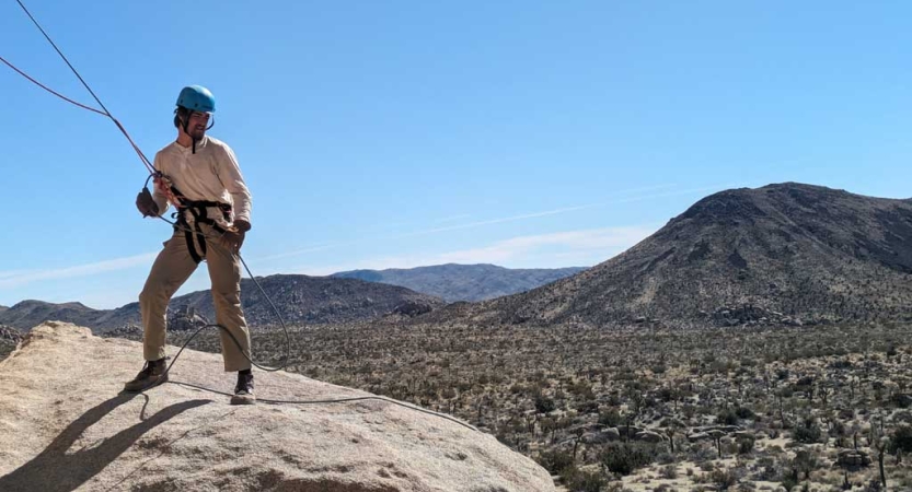 A person wearing safety gear is attached to ropes as they stand on a large boulder overlooking a desert environment. 